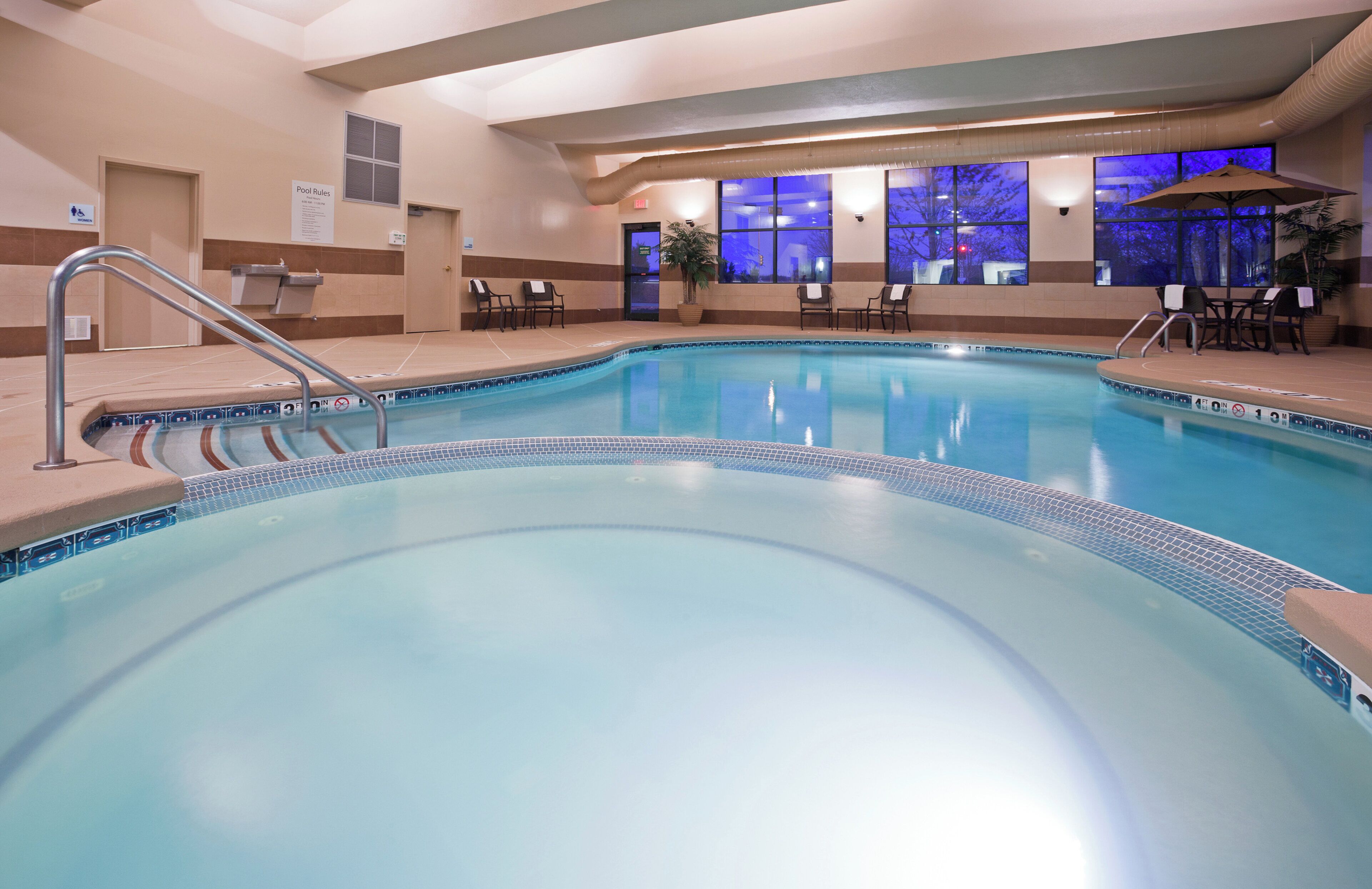 Una piscina interior con borde de azulejos, una escalera de piscina y sillas de descanso alrededor del perímetro.