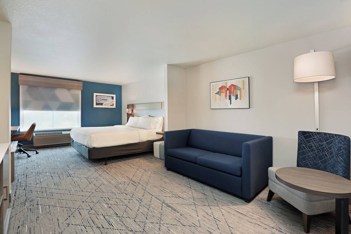 A hotel room with a bed, a blue sofa, a small round table, and a chair. There is a window with blinds, a wall-mounted lamp, and a painting on the wall.