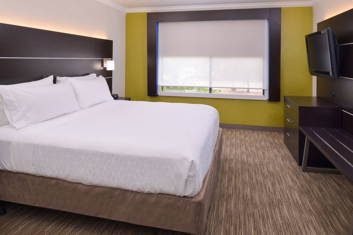 A hotel room with a large bed, a television, a window with blinds, and a desk with drawers.