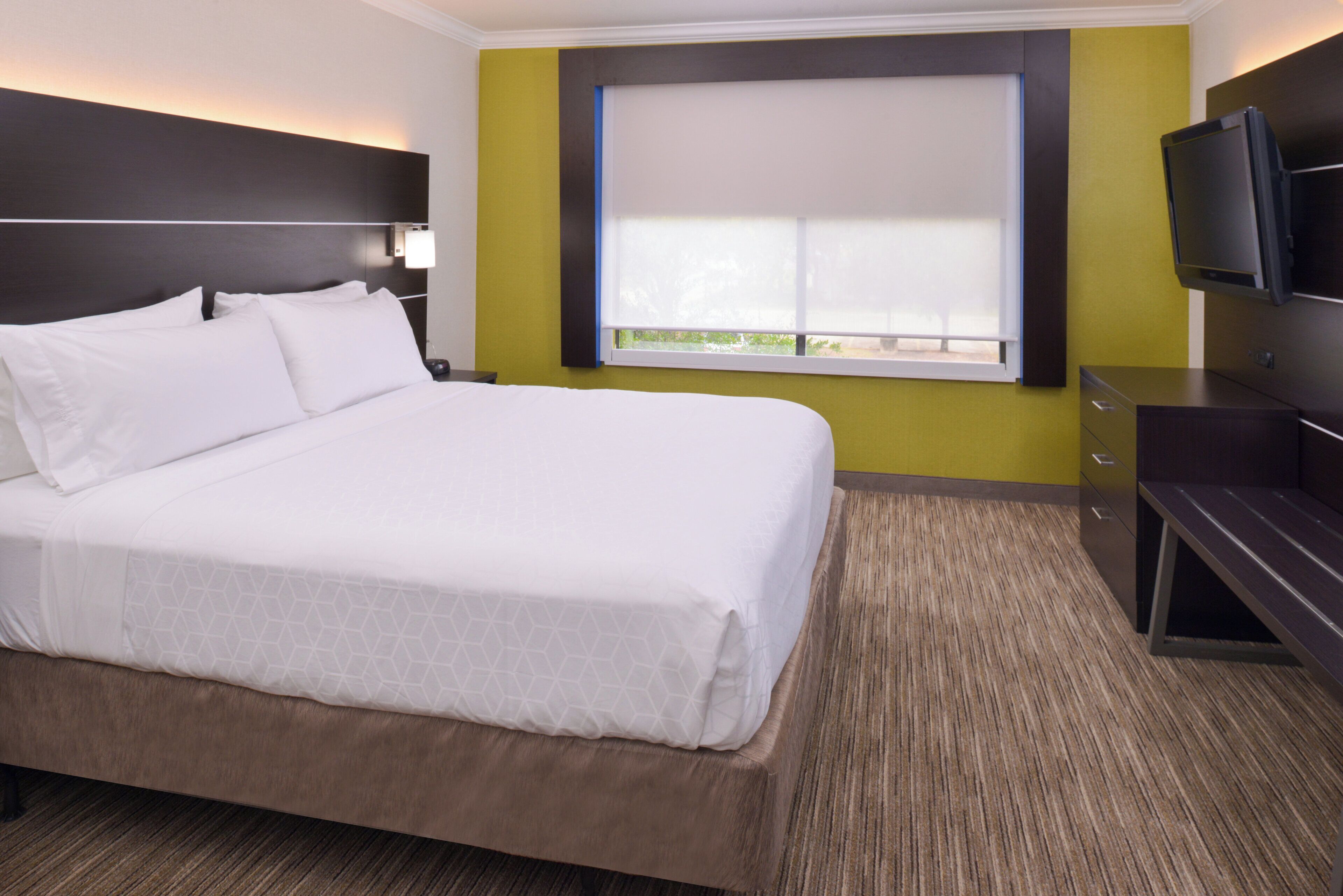 A hotel room with a large bed, a television, a window with blinds, and a desk.