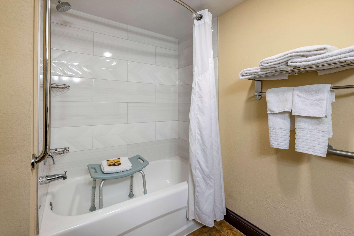 A bathroom with a bathtub, a shower curtain, a towel rack with folded towels, and a small table with a towel and a bottle.