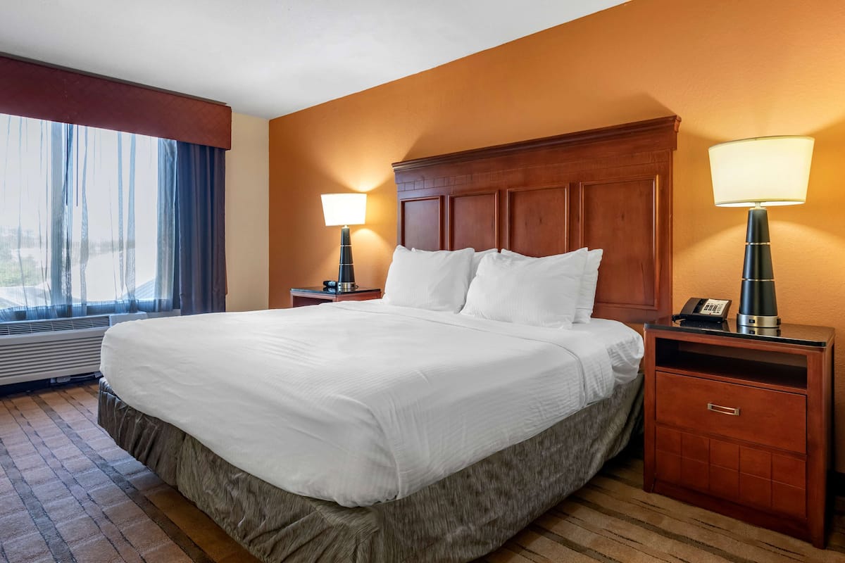 Una habitación de hotel con una cama grande, cabecera de madera, dos mesitas de noche con lámparas y un ventanal con cortinas.