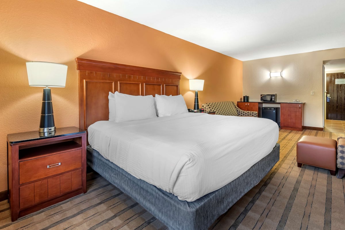 Habitación de hotel con una cama grande, una mesita de noche con lámpara, un escritorio con microondas y una kitchenette.