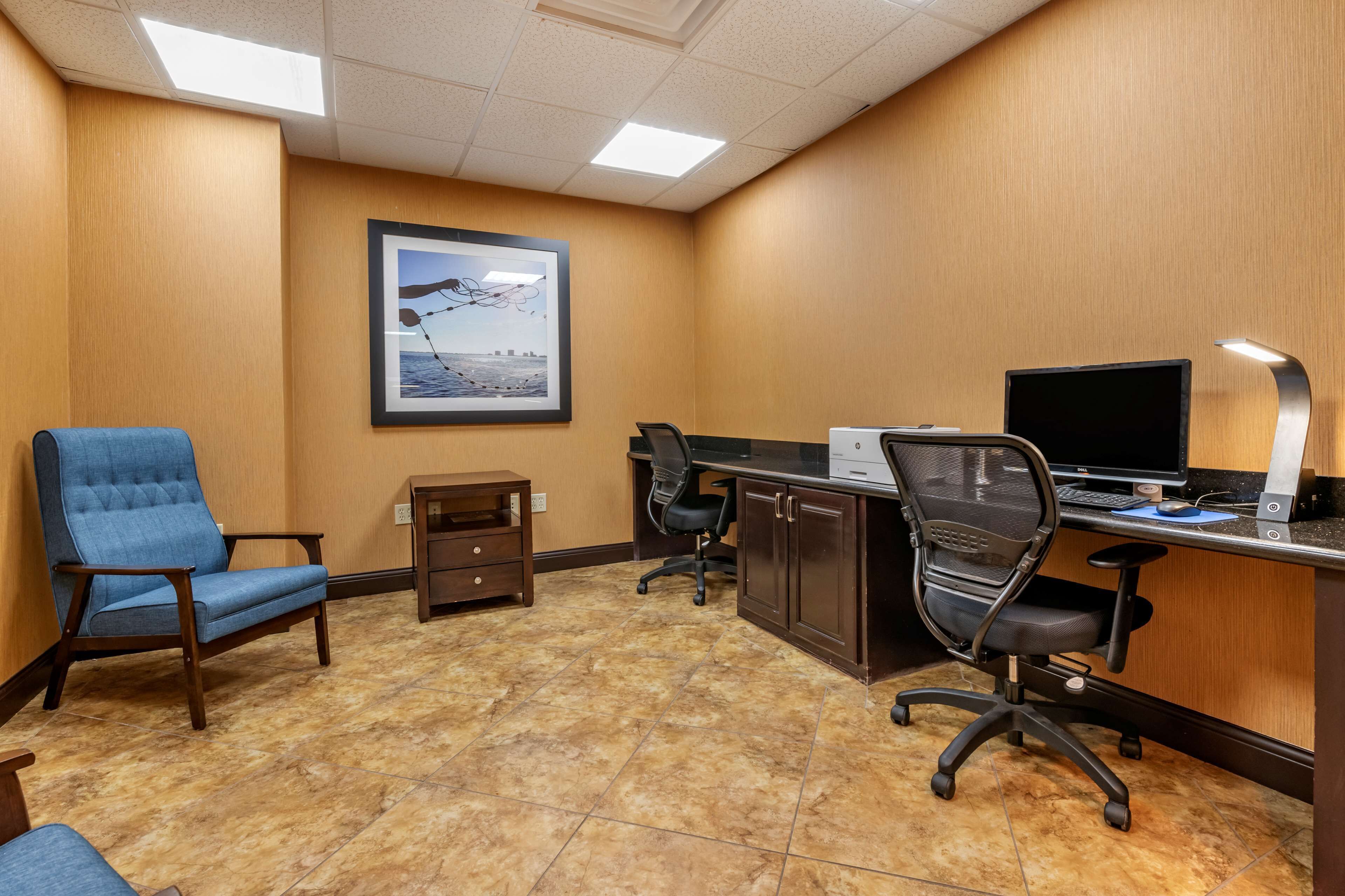 A hotel room with a wooden wall, a desk with a computer, two office chairs, a small cabinet, and a framed picture on the wall.