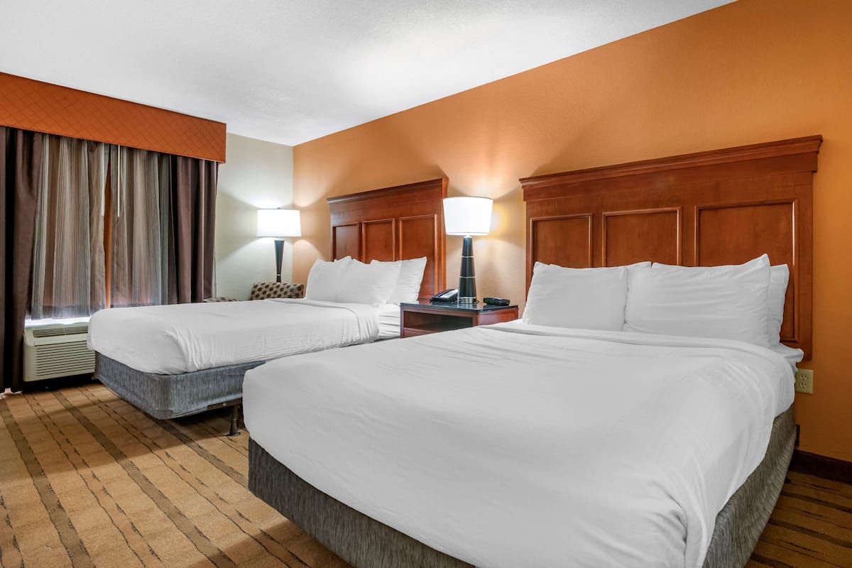 Habitación de hotel con dos camas, cabecero de madera, mesita de noche con lámpara y ventana con cortinas.