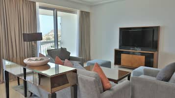 A modern living room with a glass coffee table, grey sofas, a flat-screen TV, and a view of the outdoors.