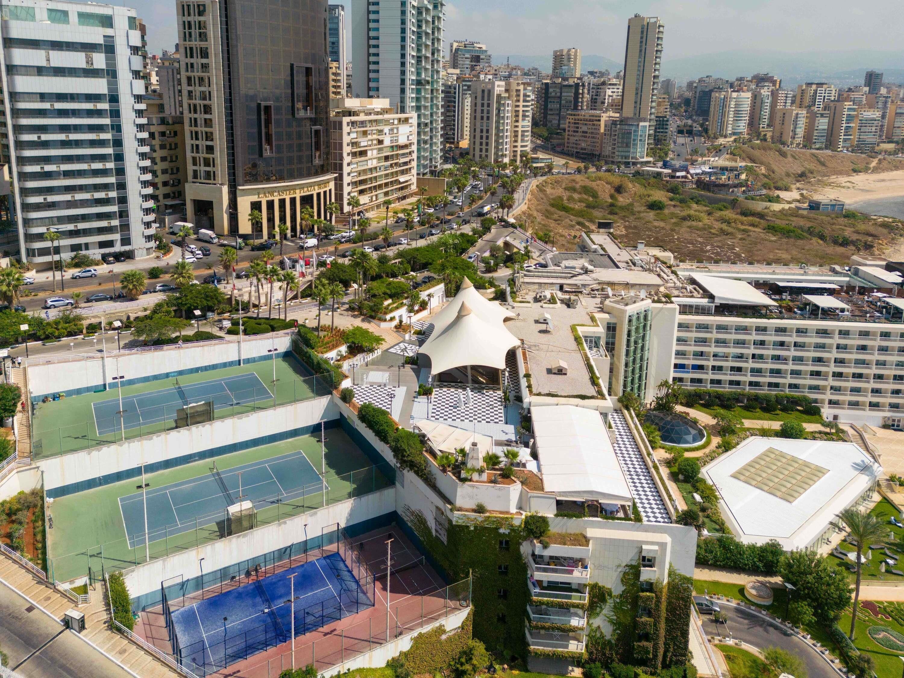 A high-rise hotel with multiple tennis courts, a rooftop garden, and a view of the city skyline.