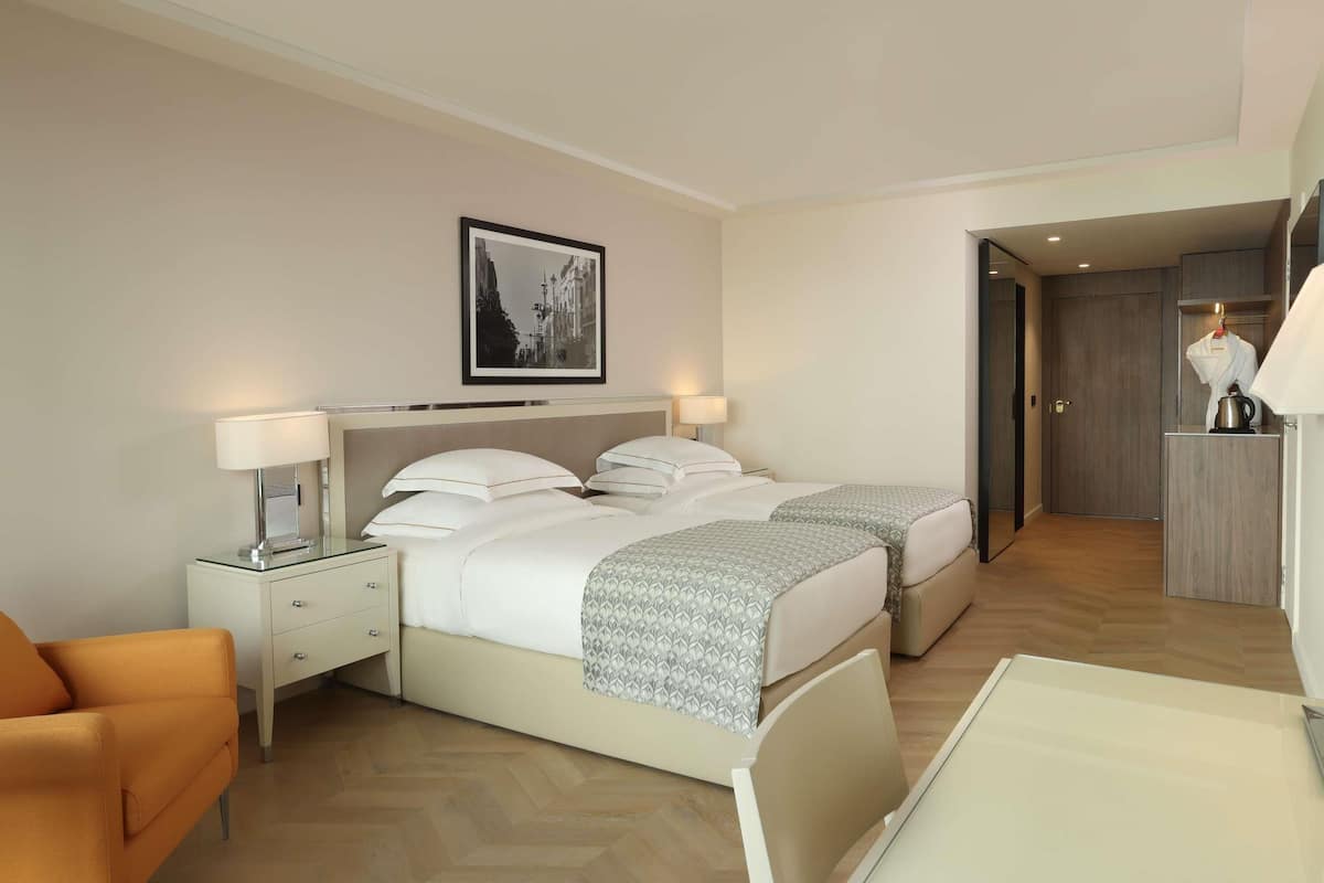 A hotel room with two beds, a nightstand with a lamp, a chair, and a framed picture on the wall.