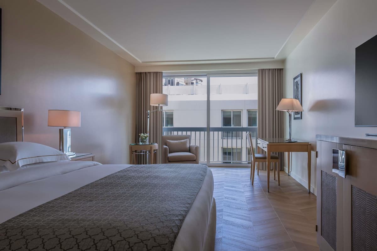 A hotel room with a bed, a desk, a chair, a lamp, and a view of the outside.