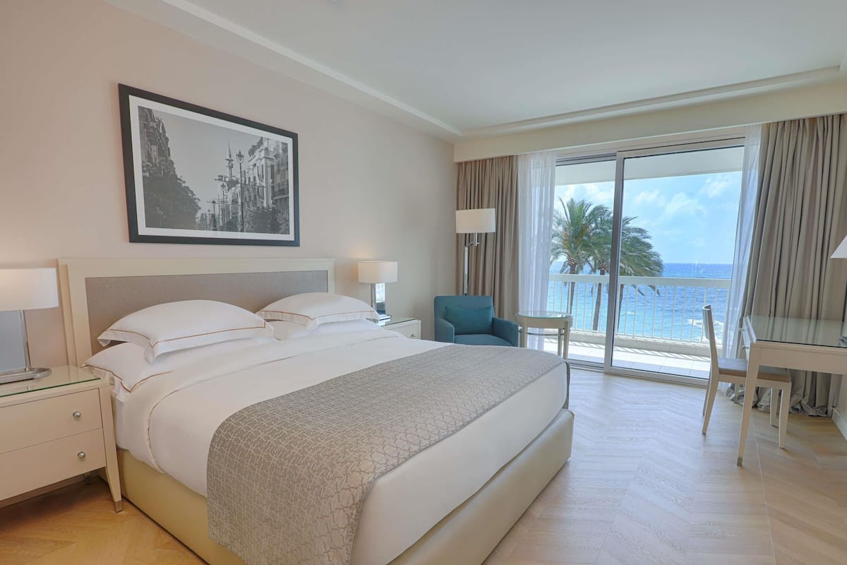A hotel room with a large bed, a desk, a chair, and a balcony with a view of the sea.
