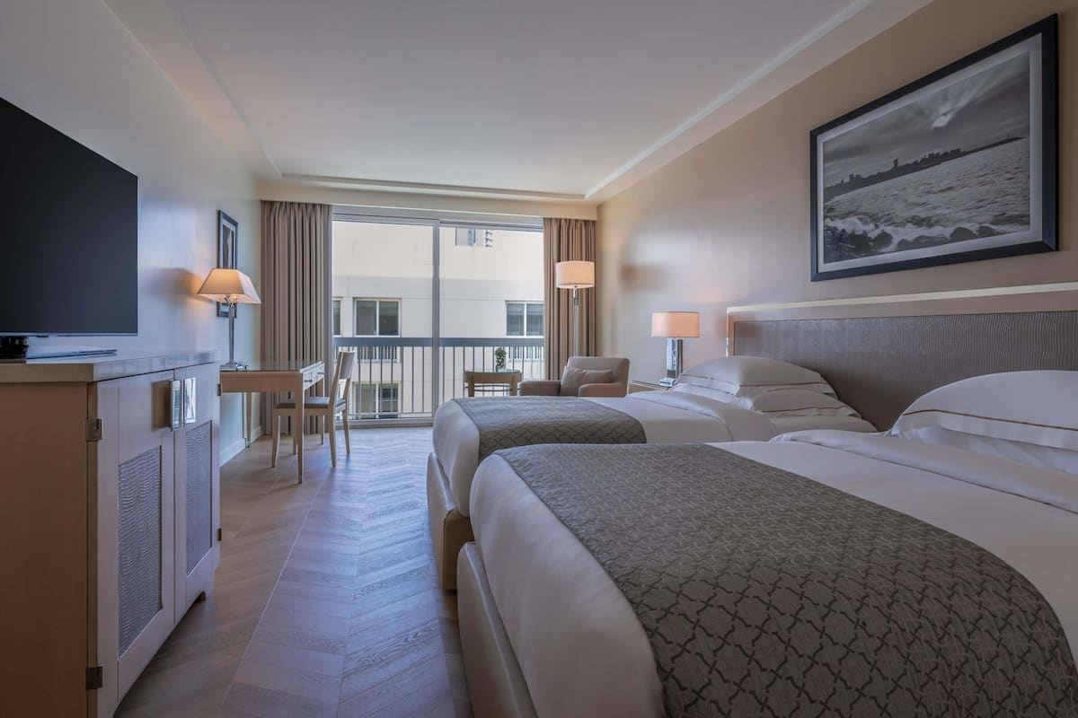 A hotel room with two beds, a TV, a desk, a chair, and a balcony with a view.