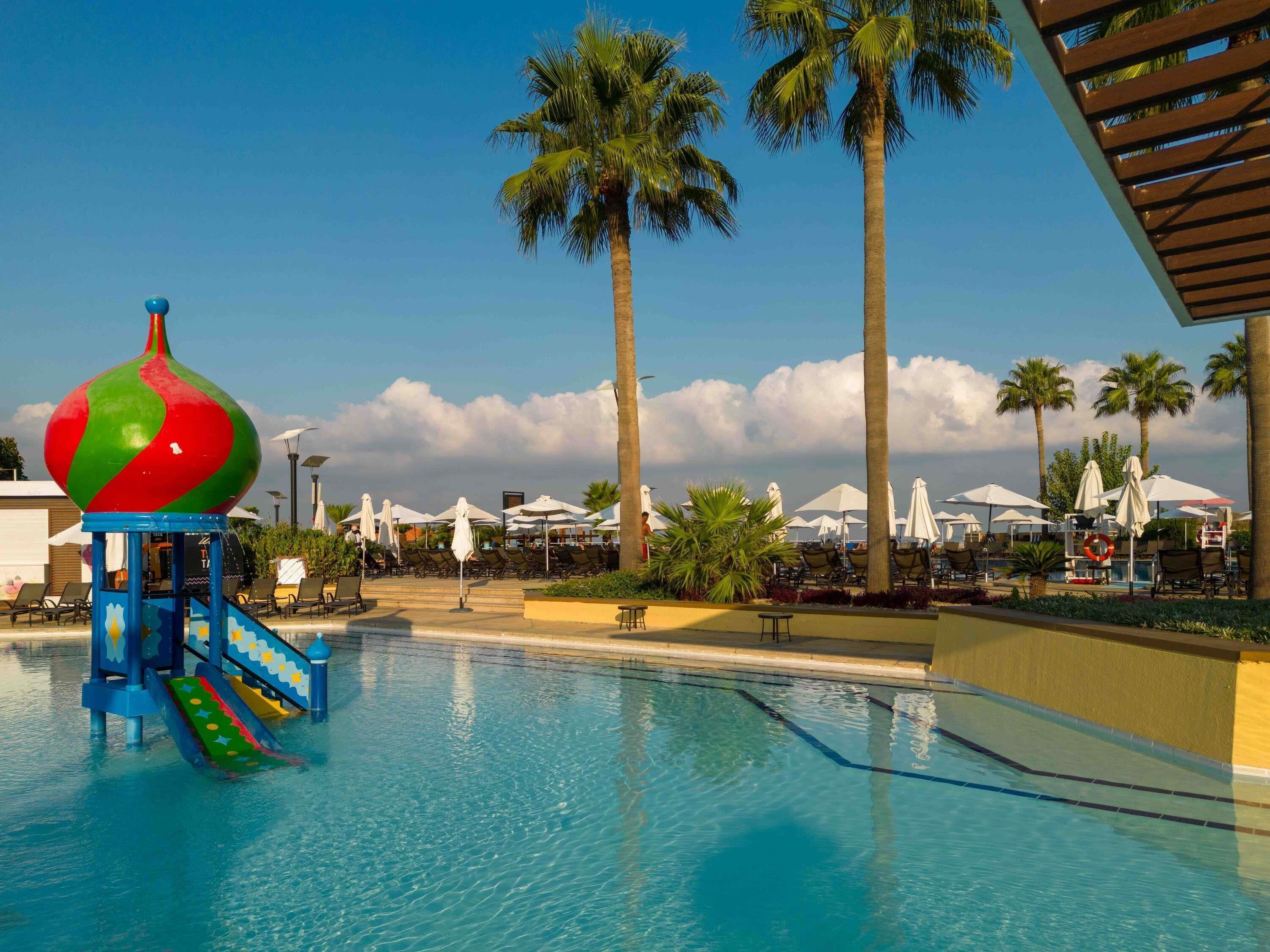 Una piscina con un tobogán acuático de colores, sillas de descanso y palmeras.