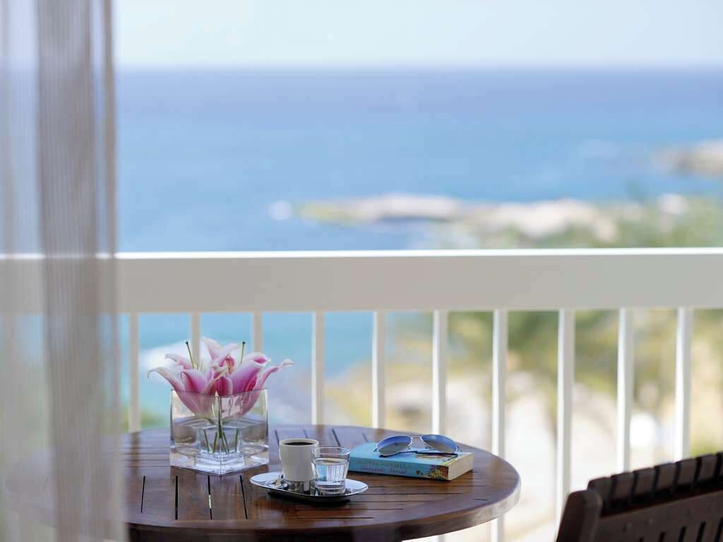 A round wooden table with a glass vase of pink flowers, a cup of coffee, and a book on a balcony overlooking the ocean.