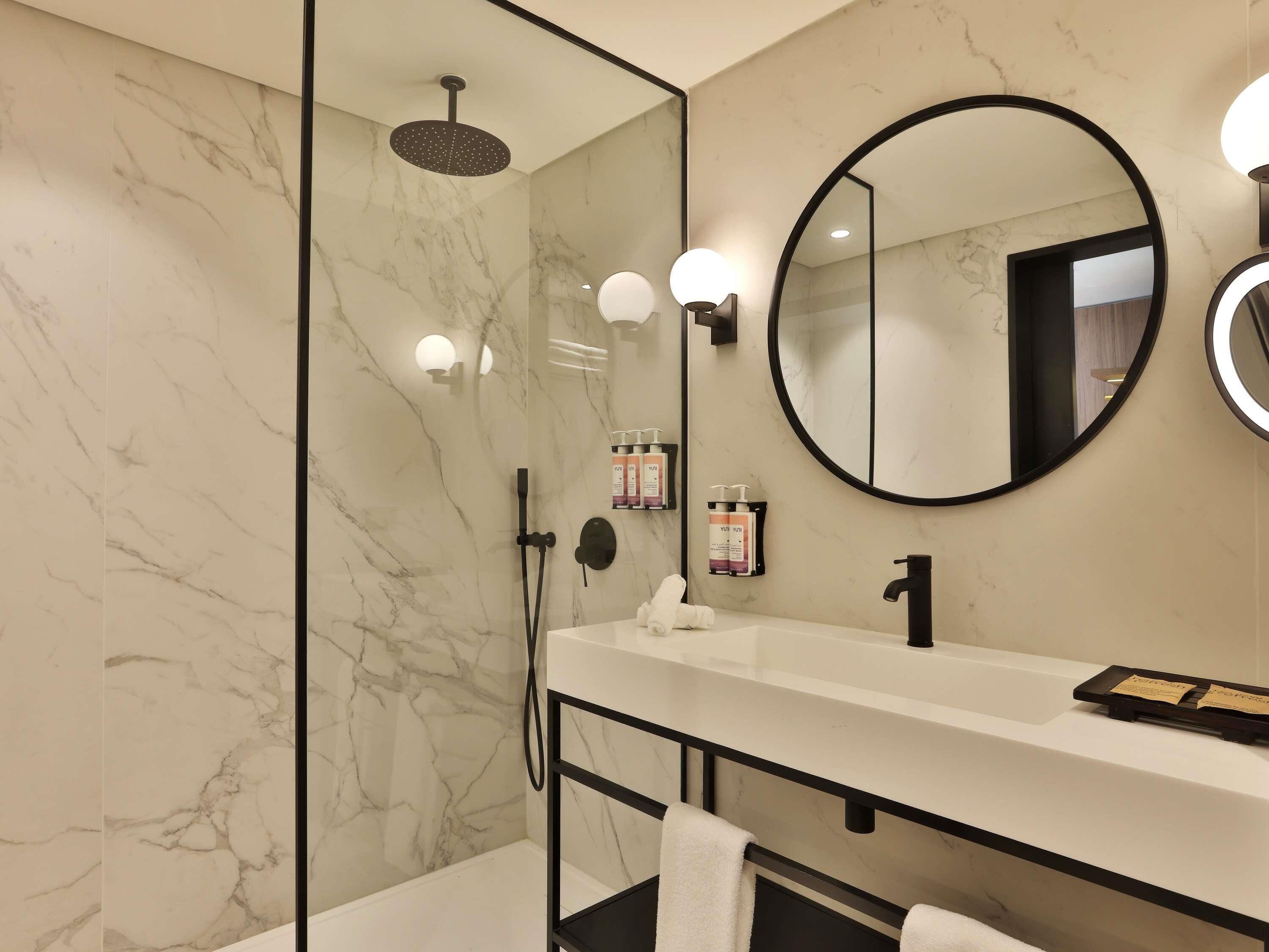 A modern bathroom with a glass-enclosed shower, a round mirror, and a white vanity with a black faucet.