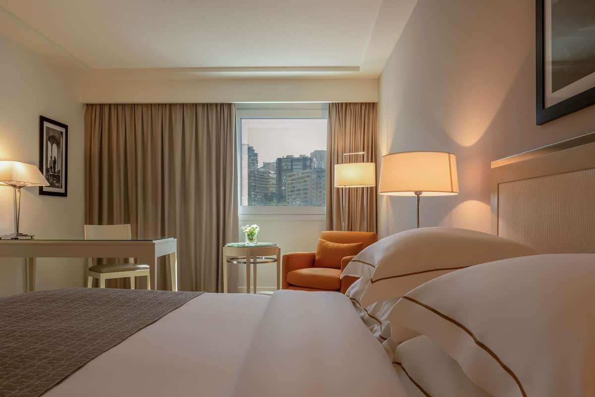 A hotel room with a large bed, a desk, an orange armchair, and a window with a city view.