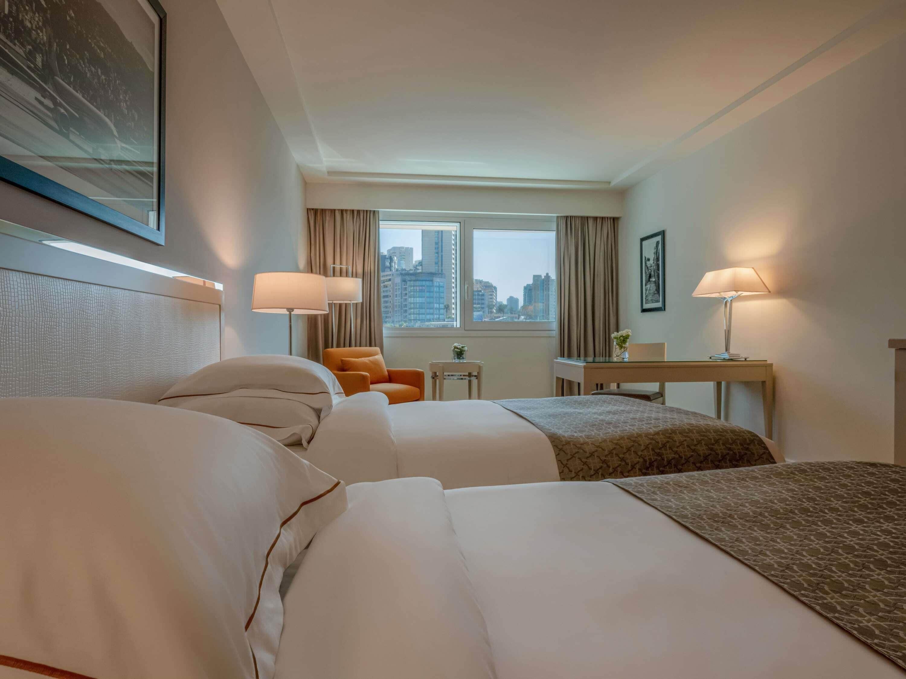A hotel room with two beds, a desk, two lamps, a chair, a small table, and a window with a city view.