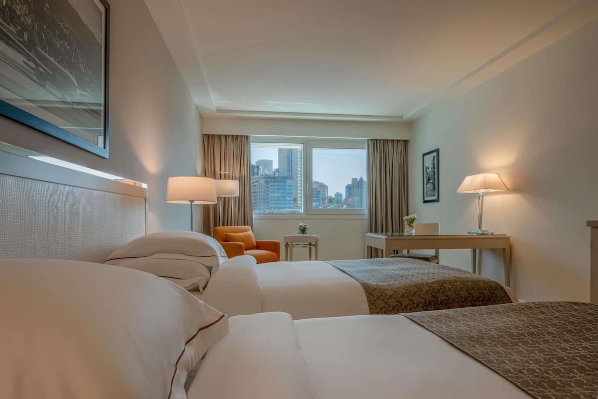 A hotel room with two beds, a desk, two lamps, a chair, a small table, and a window with a city view.