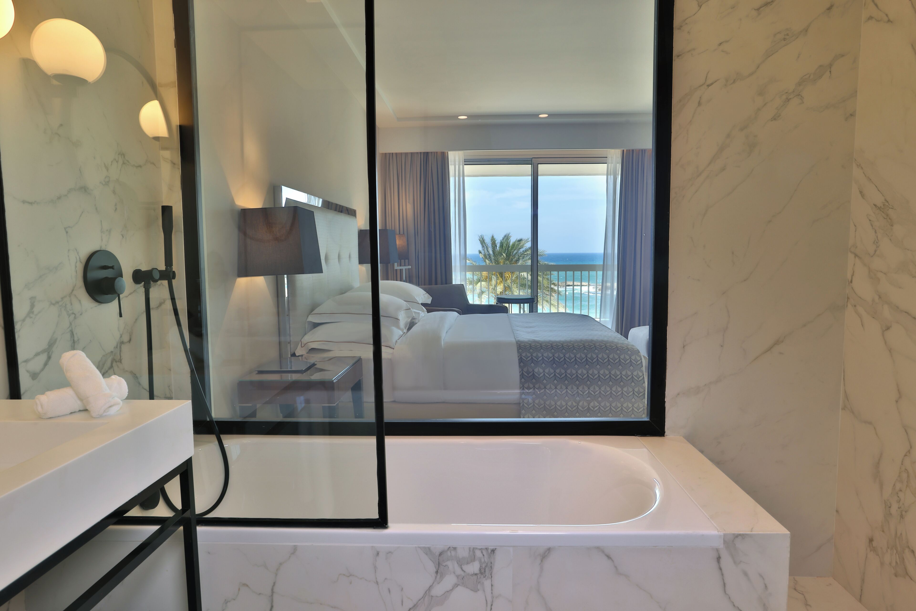 A modern bathroom with a large mirror, a bathtub, and a view of a bedroom with a balcony overlooking the sea.