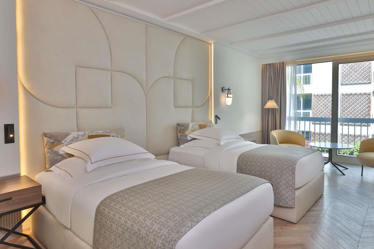 A hotel room with two beds, a small table, a chair, and a view of the outside.