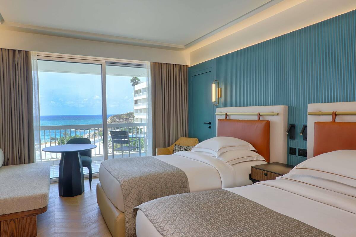 A hotel room with a balcony, two beds, a small table, and a chair.