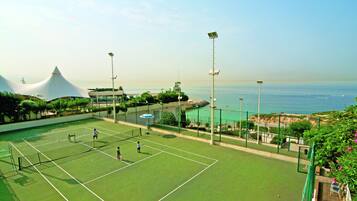 An outdoor tennis court with a net, surrounded by a fence, and overlooking a beach with a distinctive white dome structure in the background.