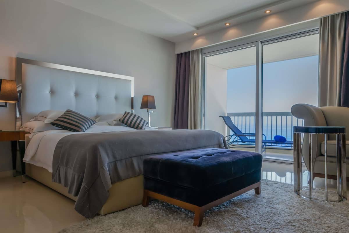 A modern bedroom with a large bed, a small ottoman, a nightstand, a lamp, and a view of the balcony.