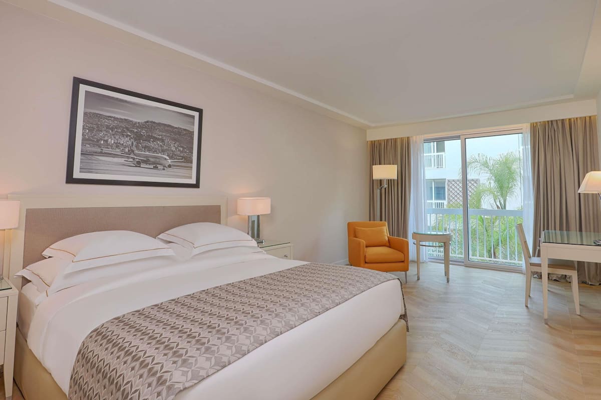 A hotel room with a large bed, two bedside tables, a desk, a chair, and a balcony with curtains.