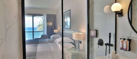 A hotel room with a large window, a bed, a bedside table with a lamp, a mirror, and toiletries on the wall.