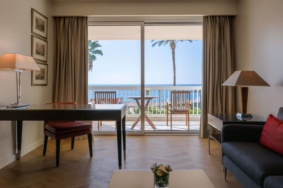 A hotel room with a balcony, a dining table, a sofa, and a desk.