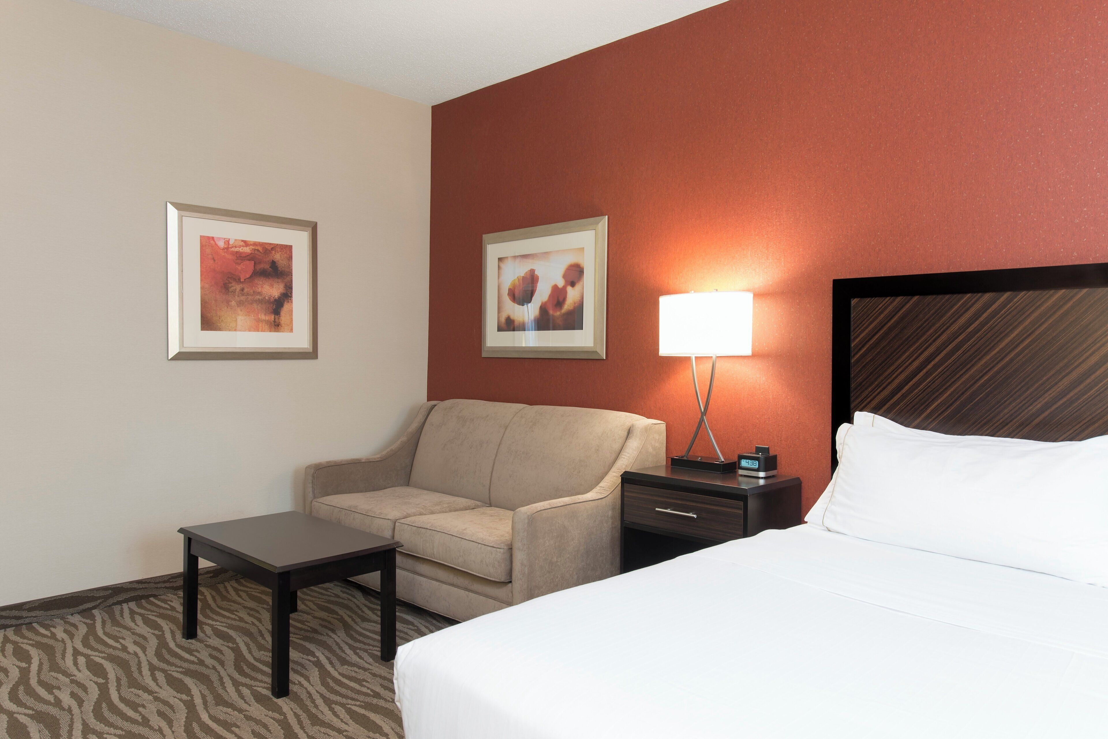 A hotel room with a bed, a chair, a nightstand, a lamp, and two framed pictures on the wall.
