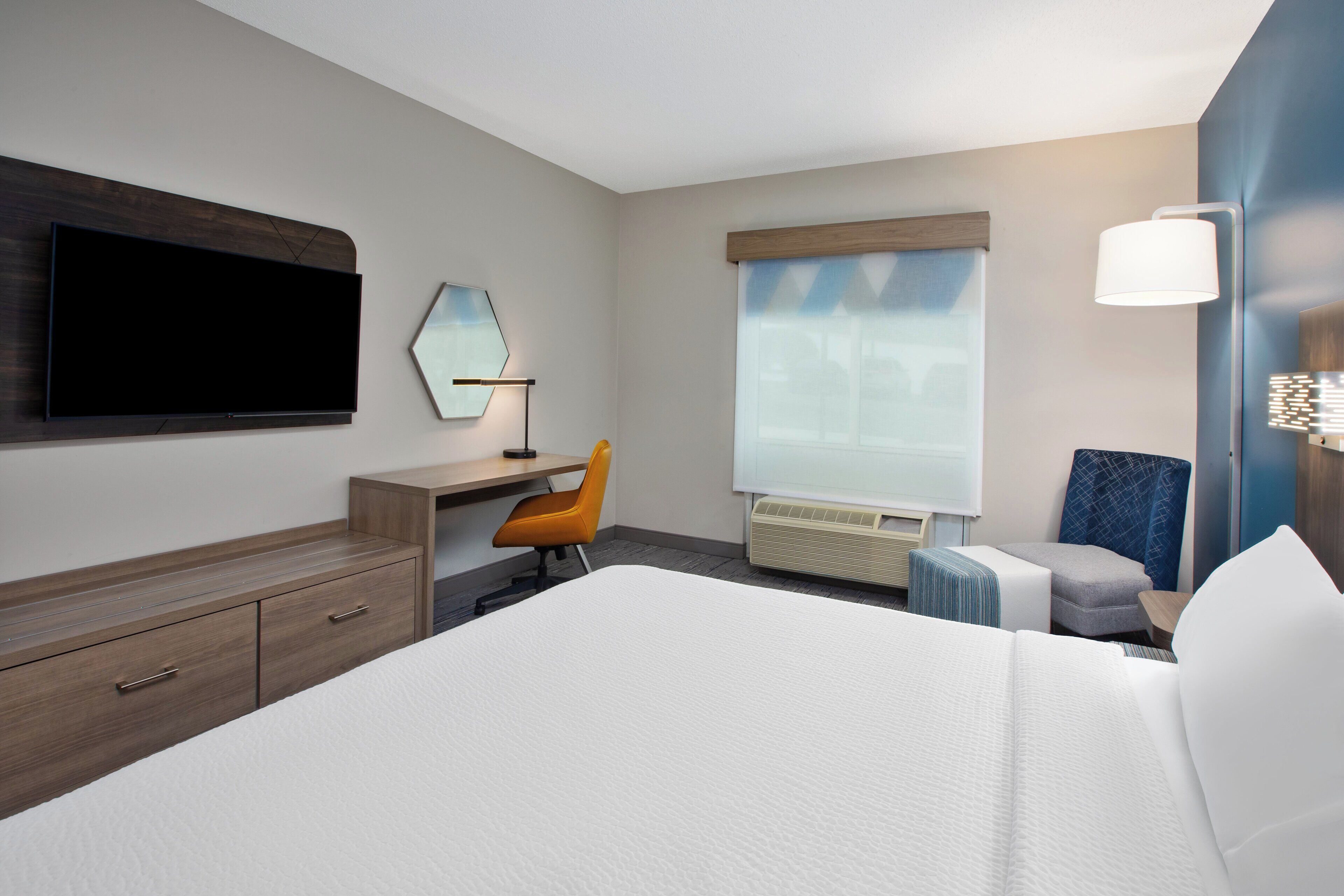 A hotel room with a large bed, a desk with a chair, a flat-screen TV mounted on the wall, a window with curtains, and a bedside table with a lamp.