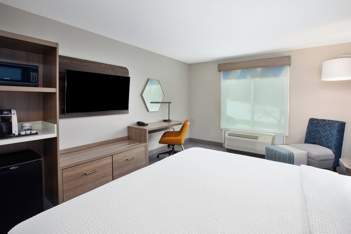 A hotel room with a large bed, a desk with a chair, a microwave, a TV, a mirror, and a window with blinds.