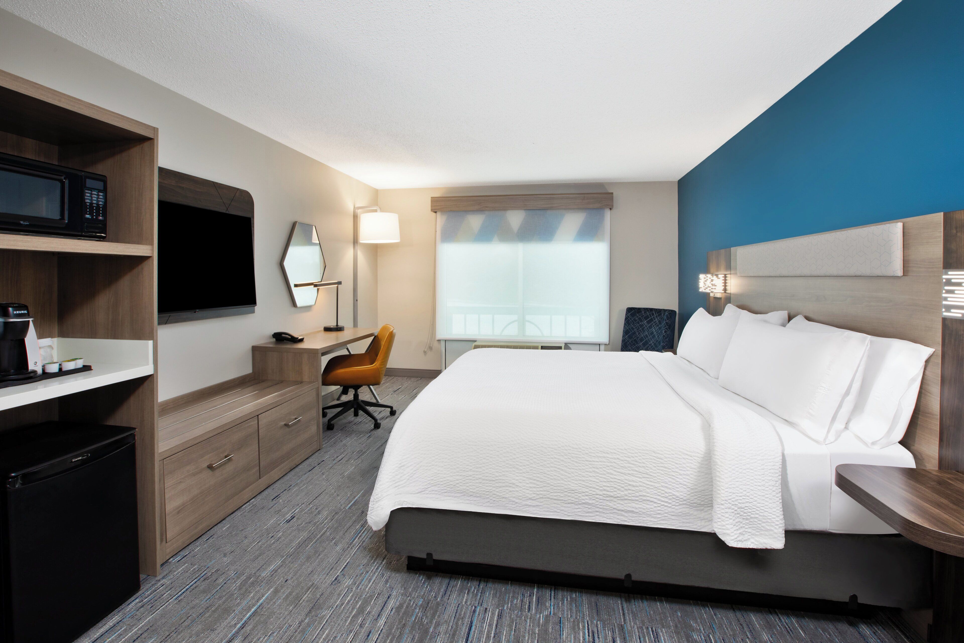 A hotel room with a bed, a desk, a chair, a TV, and a minibar.