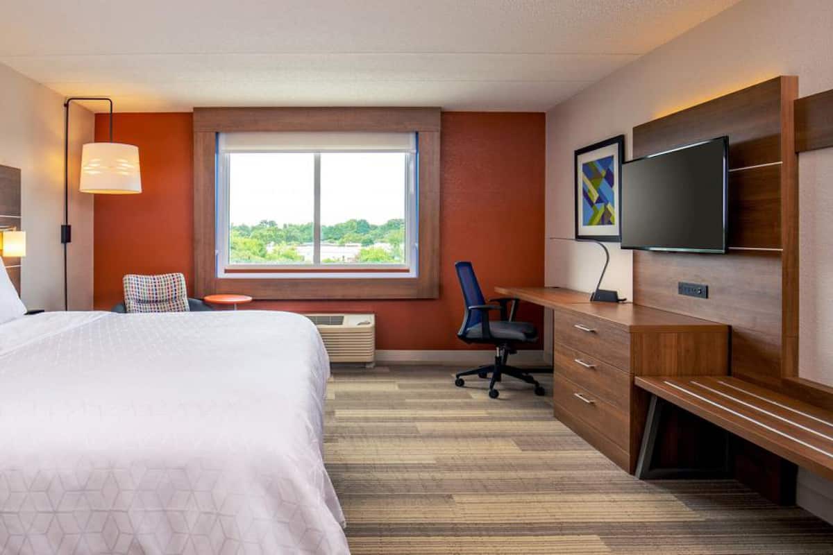 Une chambre d’hôtel avec un grand lit, un bureau, une chaise, une télévision et une fenêtre donnant sur un paysage verdoyant.