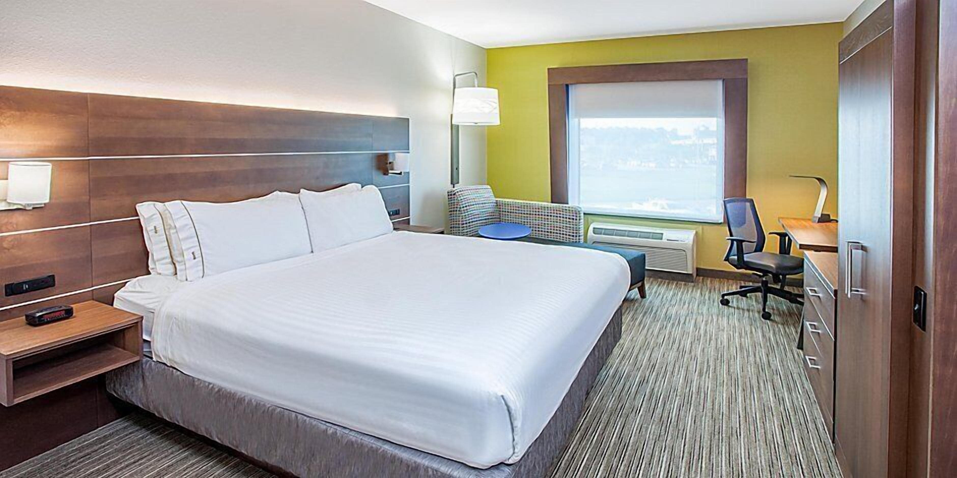 A hotel room with a large bed, a desk with a chair, a standing lamp, and a window with a view.