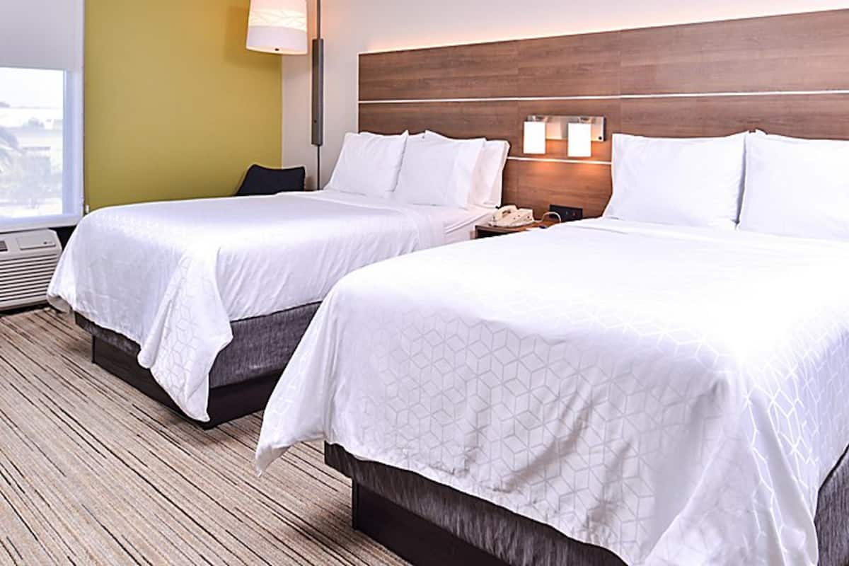 Deux lits doubles avec du linge de lit blanc dans une chambre d’hôtel, dotée d’une tête de lit en bois, d’une table de chevet avec une lampe et d’une fenêtre offrant une vue.