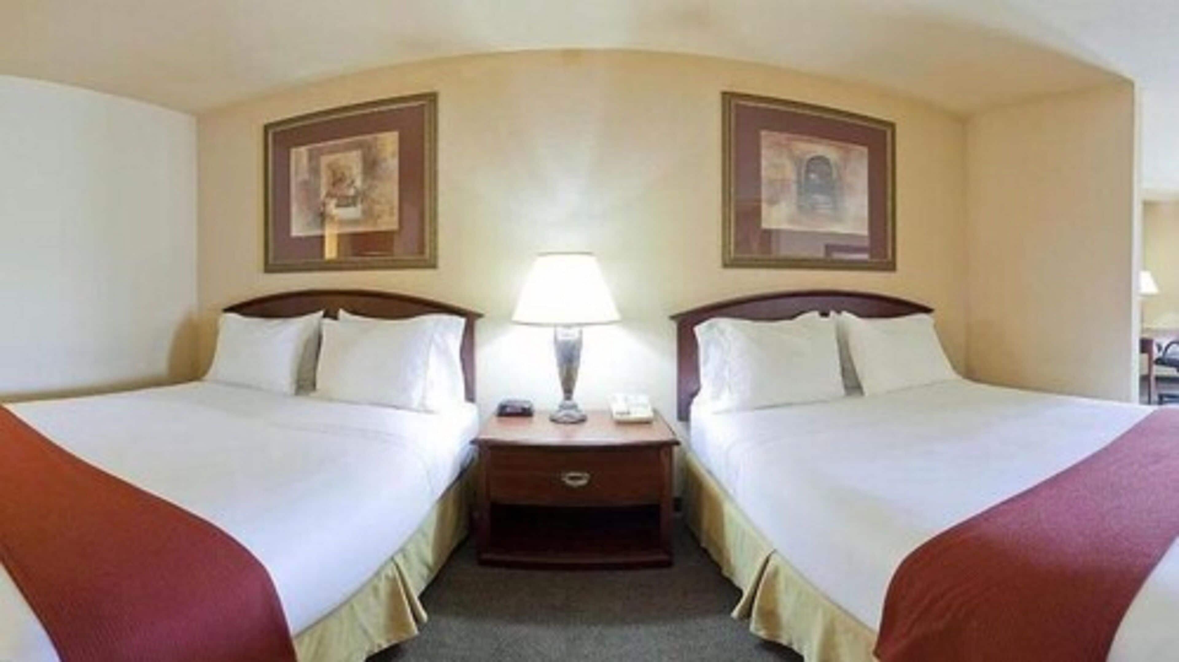 A hotel room with two beds, a nightstand with a lamp, and two framed pictures on the walls.