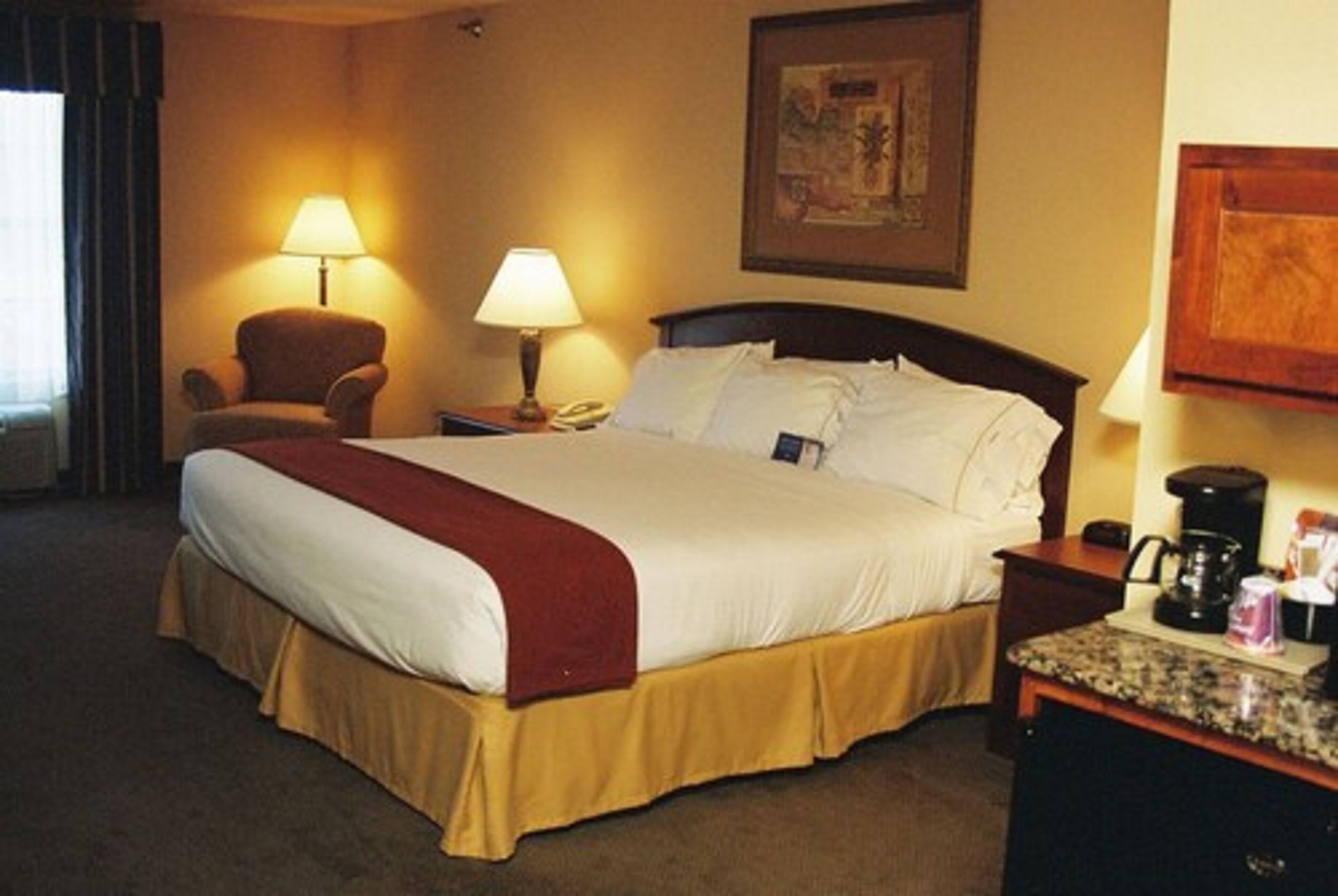 A hotel room with a large bed, two bedside lamps, a chair, a desk with a coffee maker, and a framed picture on the wall.