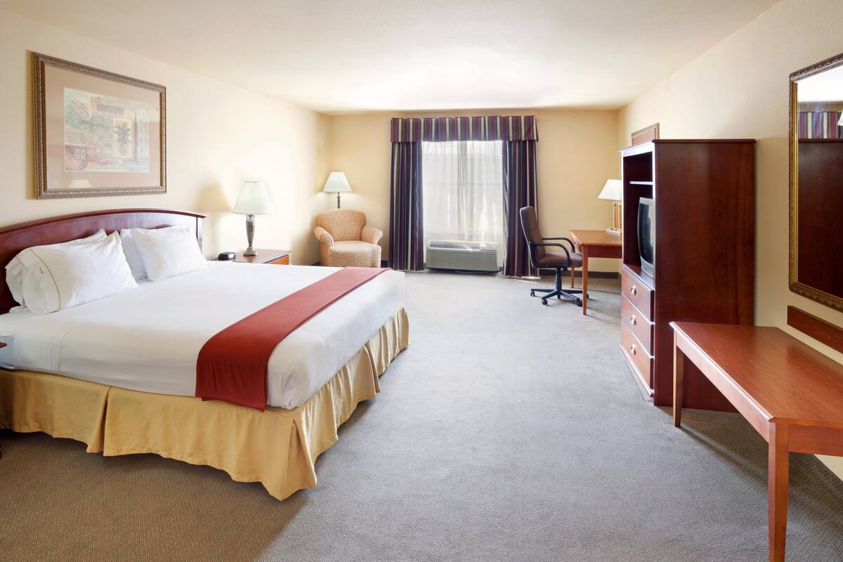 A hotel room with a large bed, a desk, a chair, a television, and an air conditioning unit.