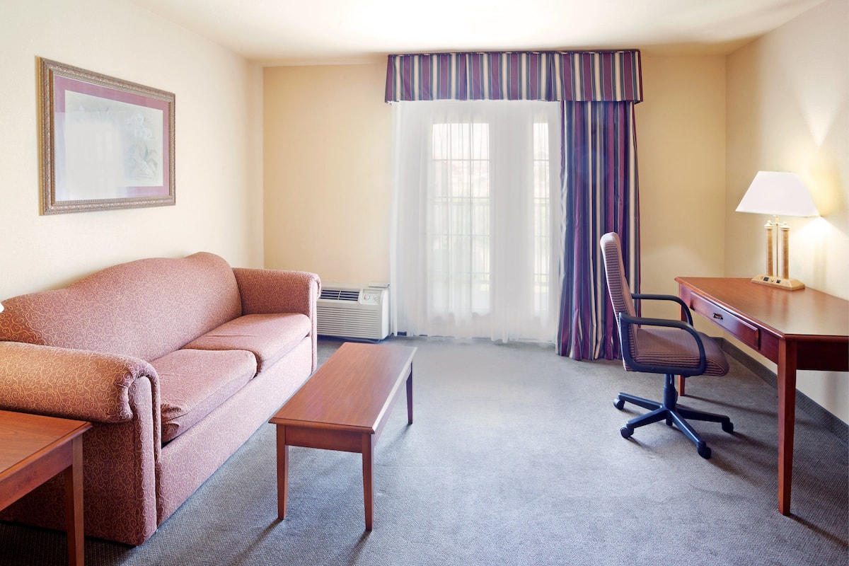 A hotel room with a sofa, a wooden desk, a chair, a lamp, and a window with curtains.