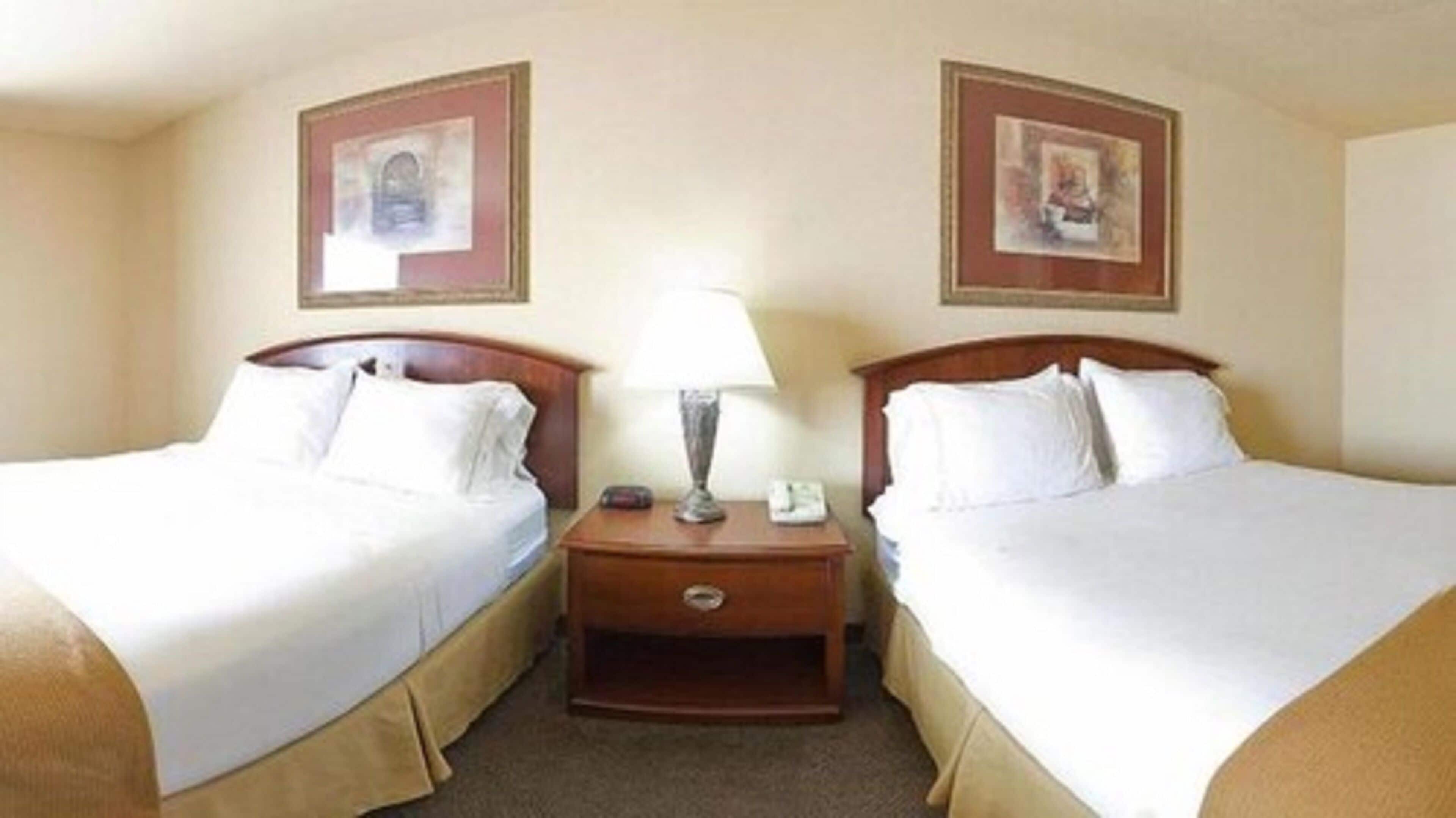 A hotel room with two single beds, a nightstand with a lamp, and framed pictures on the walls.