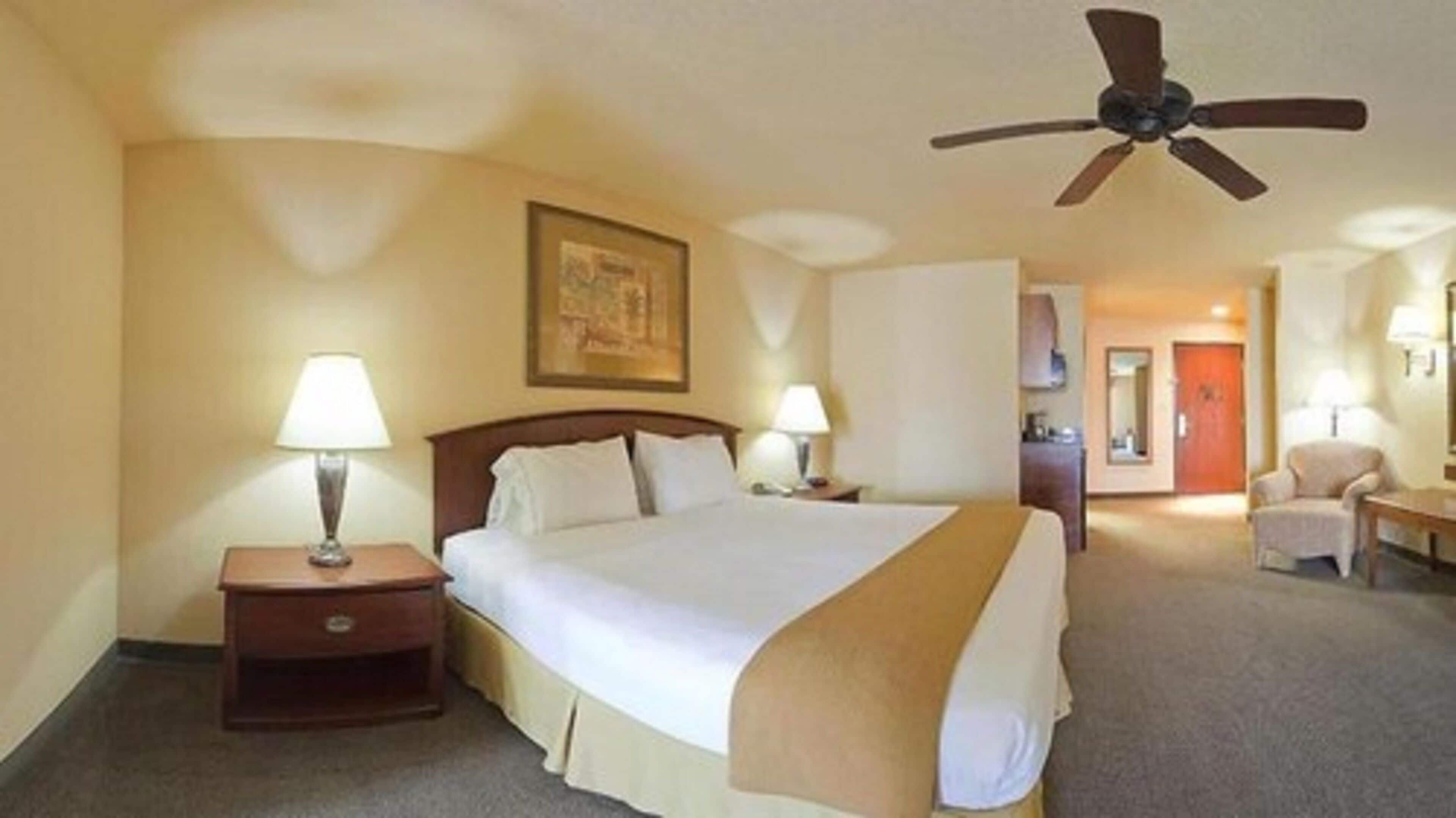A hotel room with a large bed, two bedside tables with lamps, a ceiling fan, and a door leading to a bathroom.