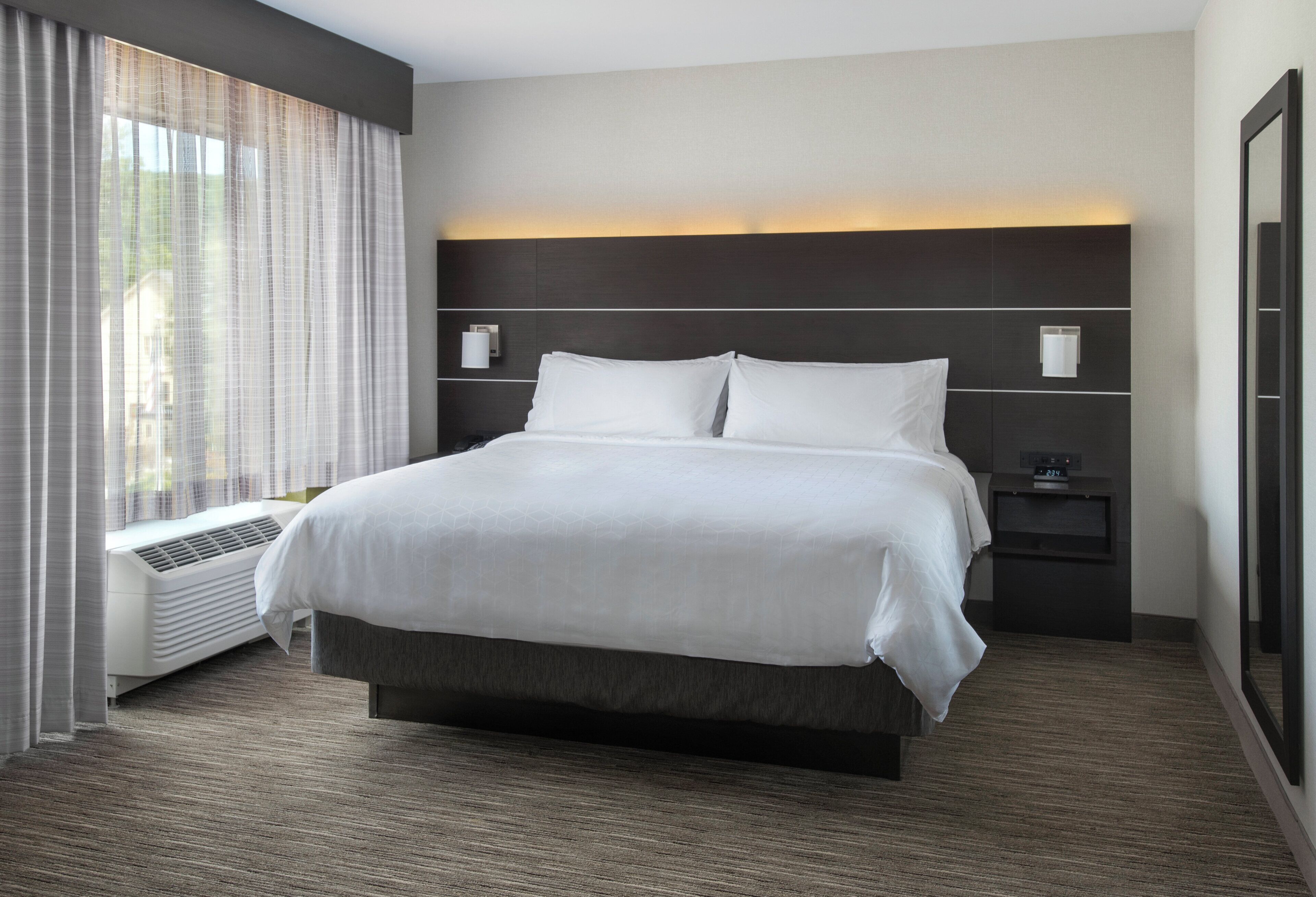 A hotel room with a large bed, a bedside table, a nightstand, a window with curtains, and a wall-mounted clock.