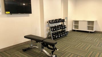 A gym with a bench, dumbbells, a television, and a white shelf unit.