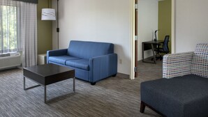 Quarto de hotel com sofá azul, uma mesa pequena, poltrona estampada e escrivaninha com cadeira.