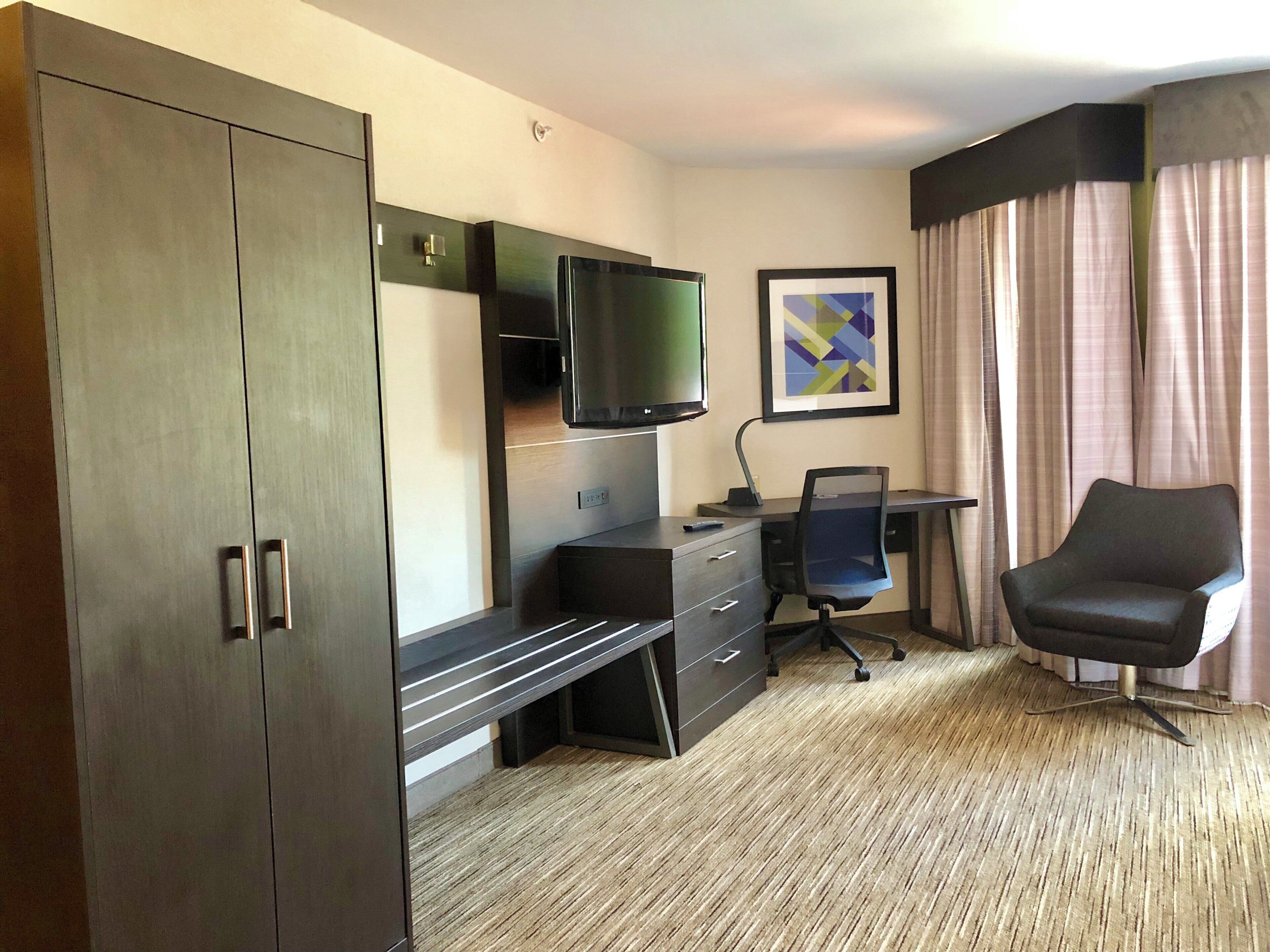 A hotel room with a large wardrobe, a desk with a chair, a television, and a framed picture on the wall.