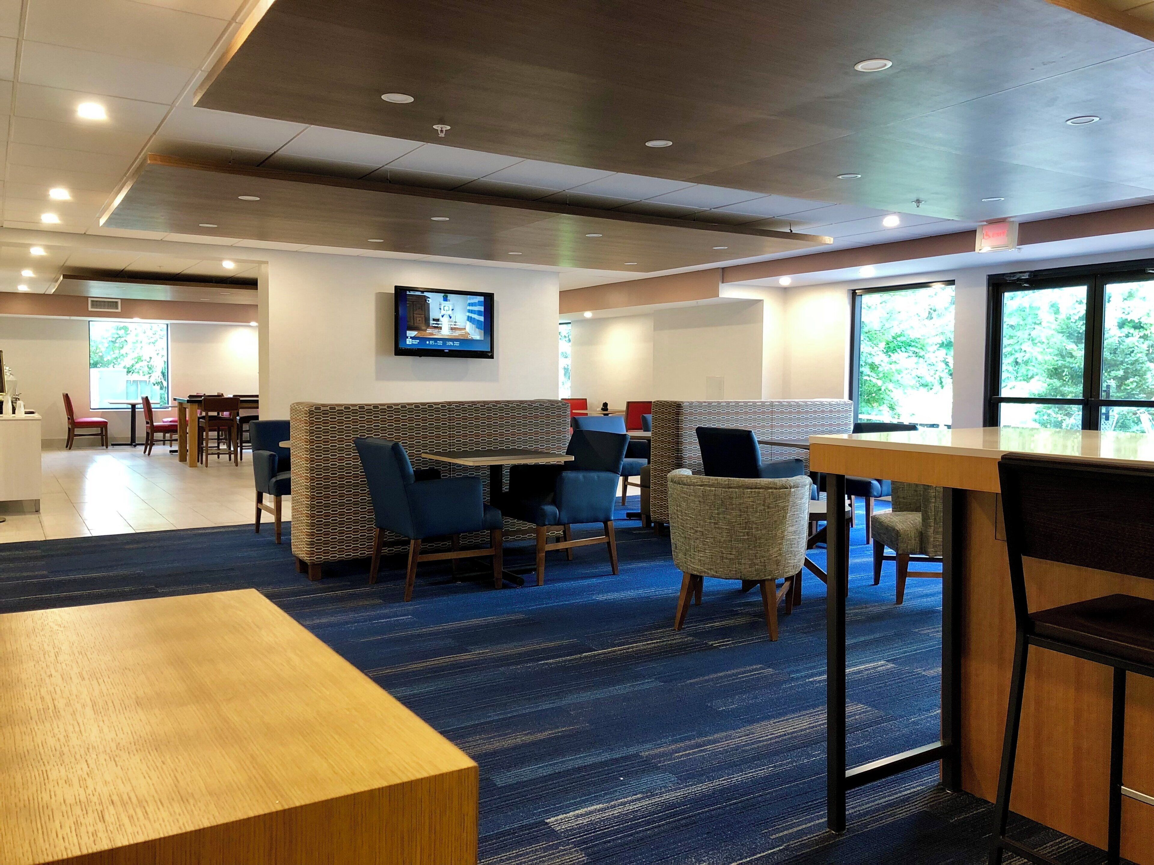 A hotel lobby with seating areas, a television mounted on the wall, and large windows providing natural light.