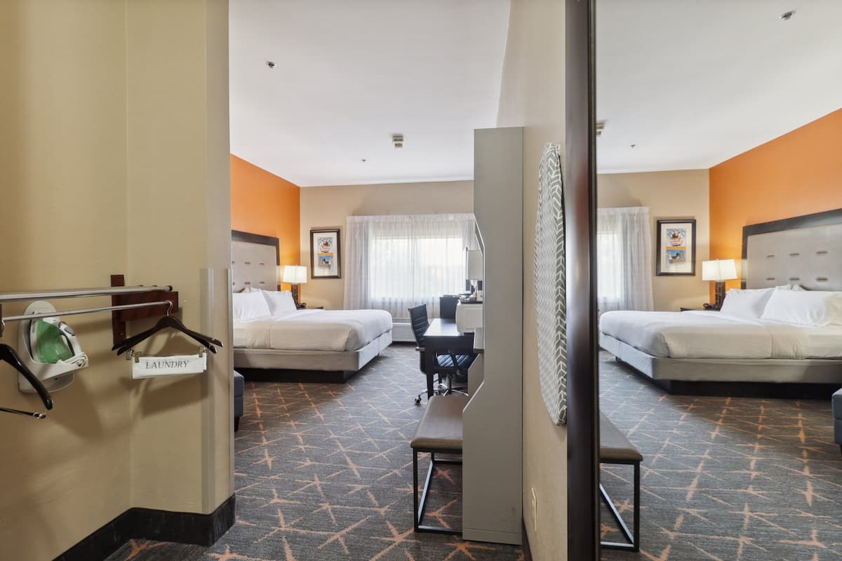 A hotel room with two beds, a desk, a chair, a bench, a bathroom with a toilet and a sink, and a mirror.