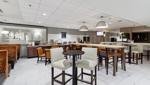 A modern hotel lobby with a bar area, seating arrangements, and a visible kitchen in the background.