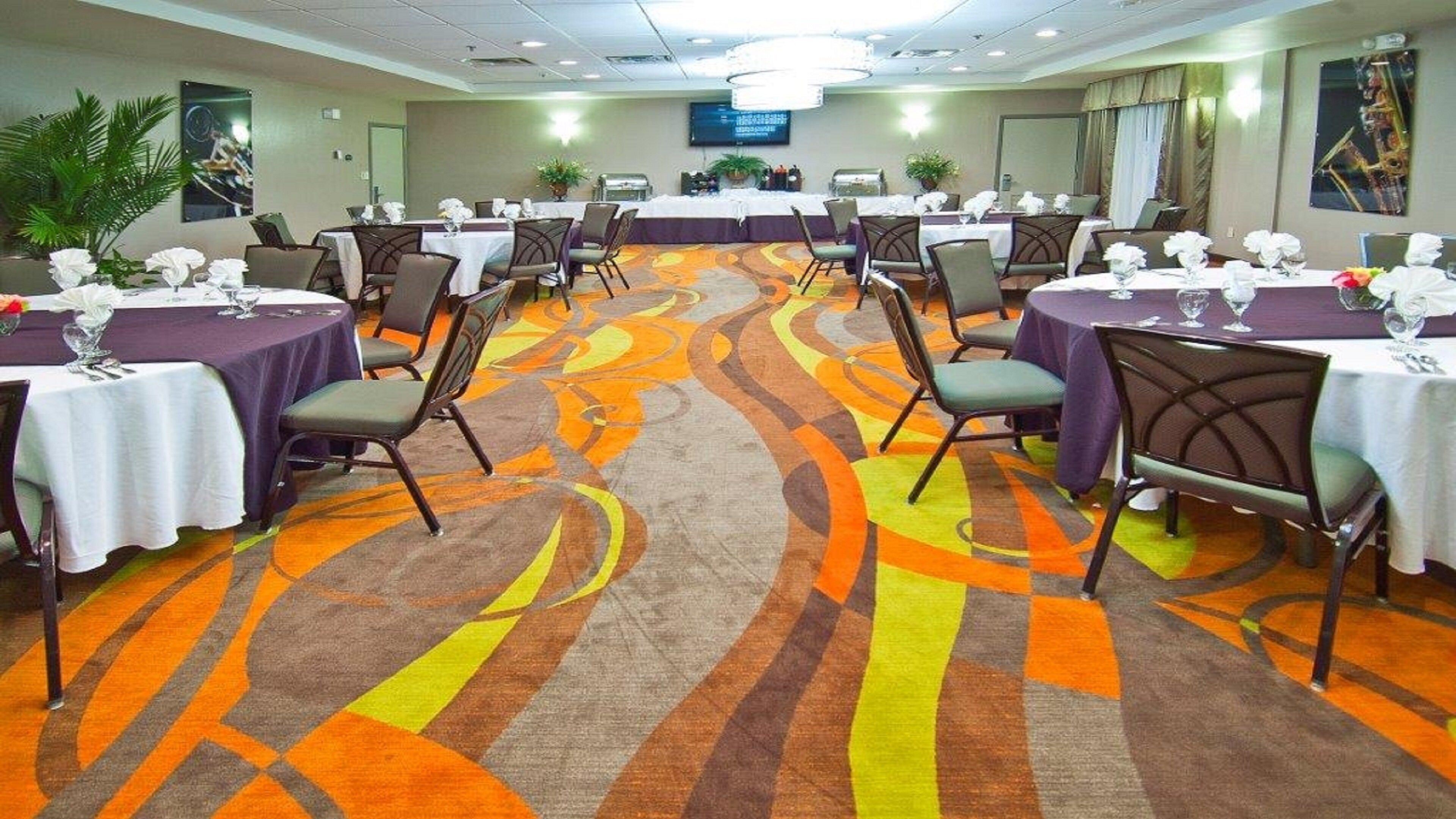 A conference room with multiple tables and chairs, a patterned carpet, and a television screen on the wall.